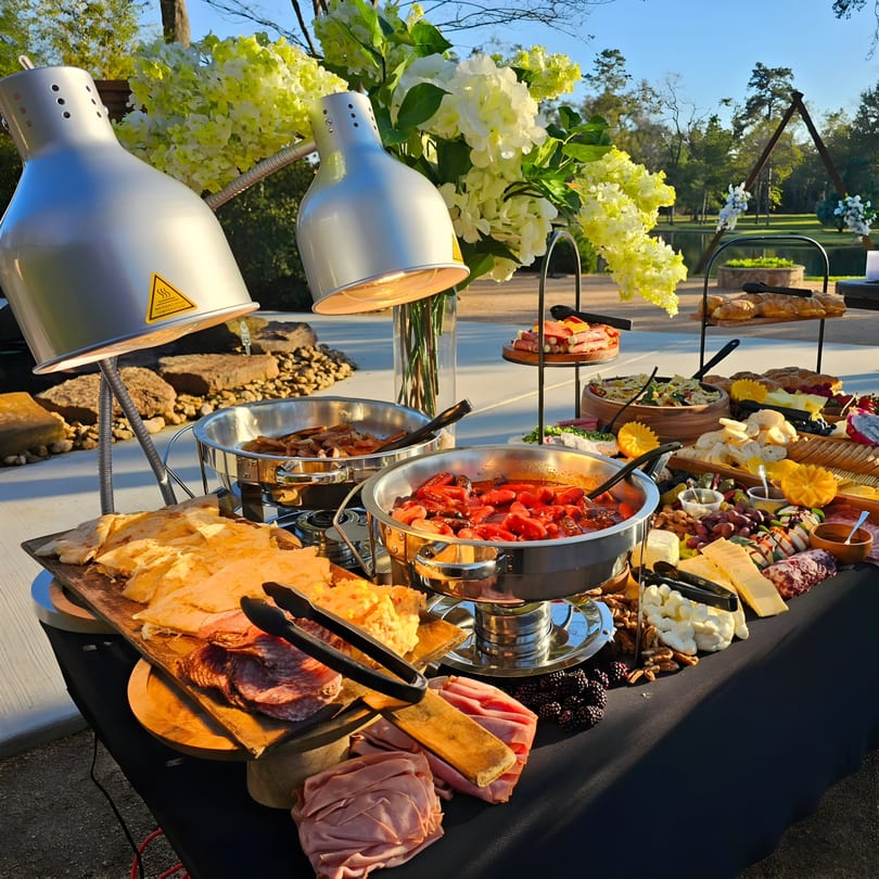 An outdoor buffet table laden with Mexican food, cheeses, meats, and fruit under heat lamps.