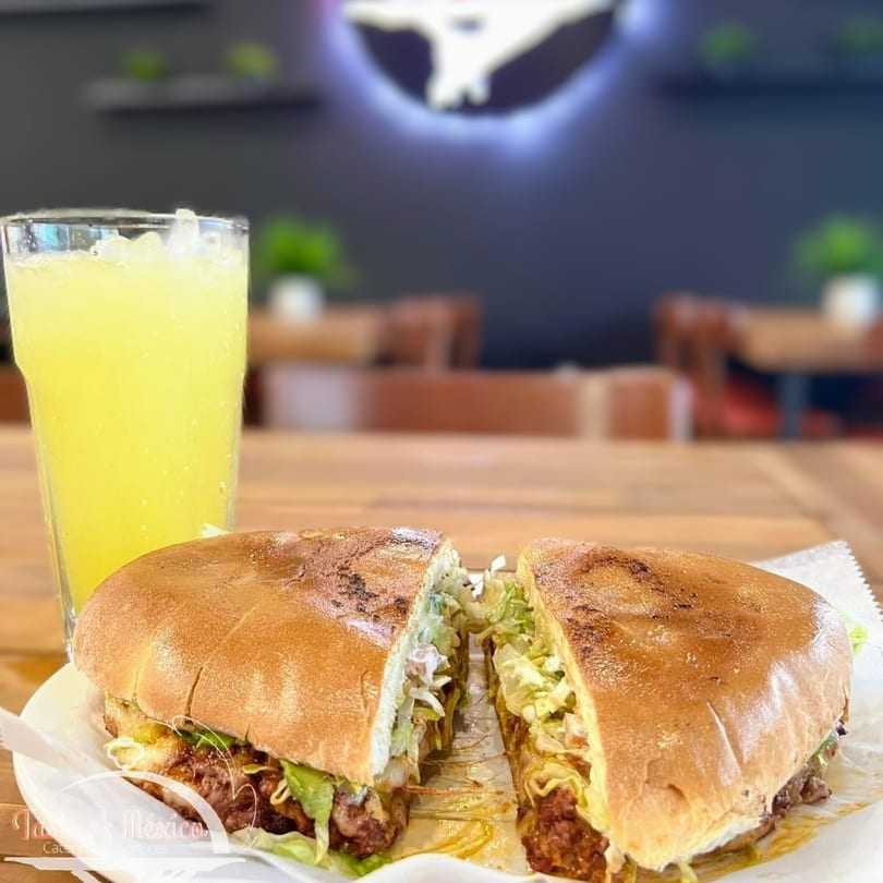 A Mexican torta sandwich, cut in half, with a glass of iced lemonade on a wooden table.