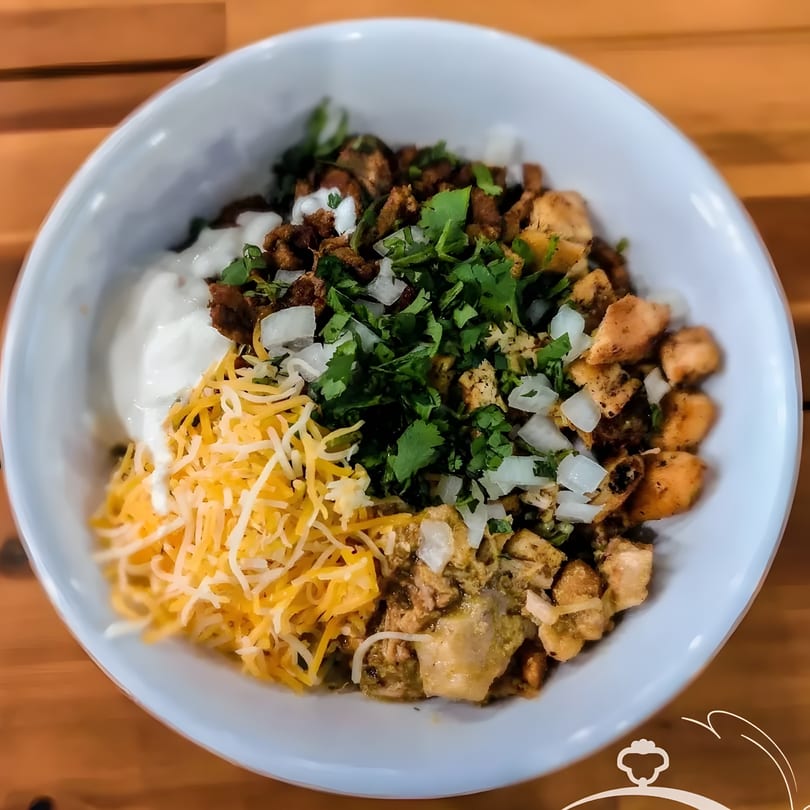 A Mexican bowl with meat, cheese, sour cream, cilantro, and onions on a wooden table.