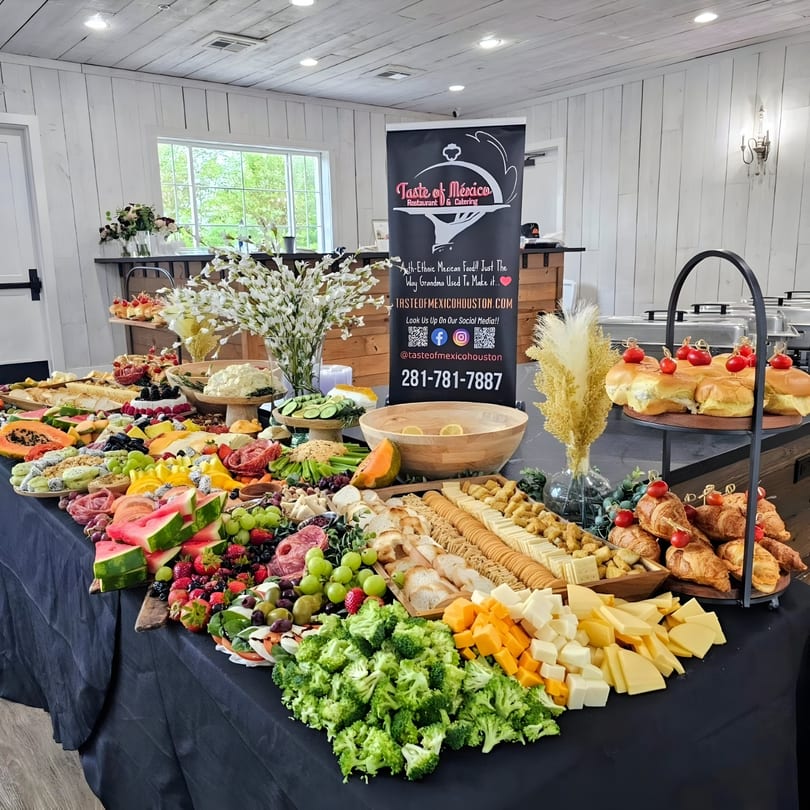 A large buffet table laden with a variety of fruits, cheeses, meats, and pastries, with a "Taste of Mexico" banner.