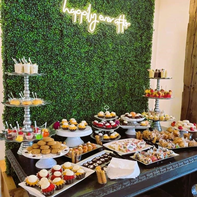 A dessert table with various pastries, cupcakes, and cookies against a green wall with a neon sign.